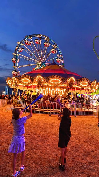 Two kids hold light-up blue wands in front of the illuminated ferris wheel and carousel at Old Canal Days 2026 as the sky fades to dusk