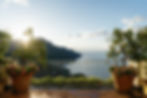 Sun-drenched terrace at Belmond Caruso in Ravello with two potted lemon trees and terracotta planters of pink flowers flanking a white iron balustrade, with the Amalfi Coast mountains and blue Tyrrhenian Sea glowing in the morning light.