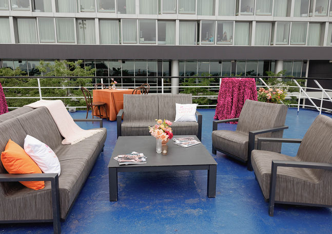 Outdoor lounge area on the upper deck of the Spirit of Chicago featuring grey modular seating, a low coffee table with a floral centerpiece, orange accent pillows, and fuchsia textured linens in the background with the Chicago skyline visible beyond the railing.