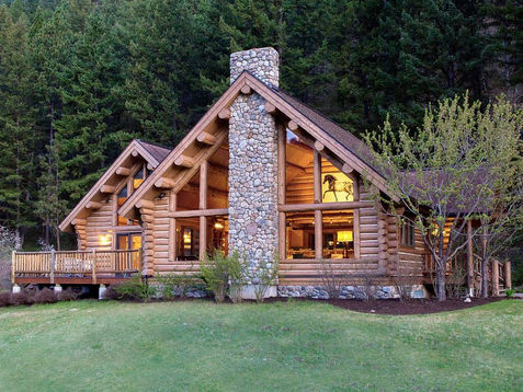 Exterior of a luxury log cabin at Triple Creek Ranch in Montana, nestled in pine forest with a stone fireplace chimney and warm interior lighting