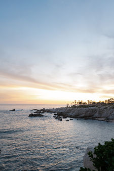 Sunset landscape of the Punta Ballena coastline at Esperanza Resort in Cabo San Lucas, showing the dramatic white volcanic cliffs, jagged rocky outcroppings emerging from the calm Sea of Cortez, a narrow sandy cove at the base of the cliffs, and the silhouetted palm-lined headland glowing gold against a soft pastel dusk sky.