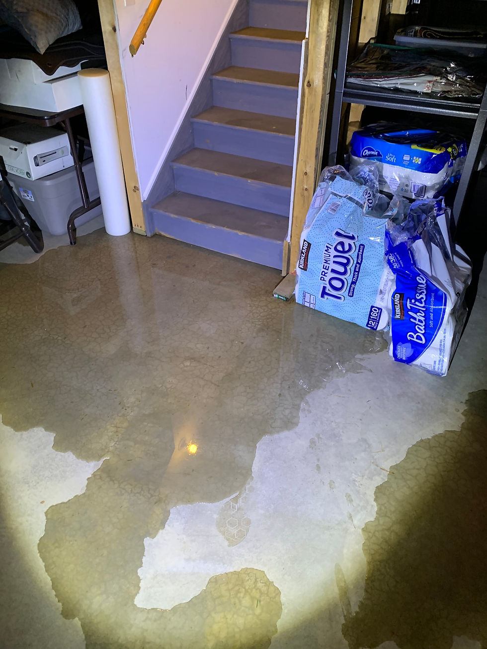 Standing water on basement floor from floor seepage near staircase, saturating stored household goods including paper towels and bath tissue