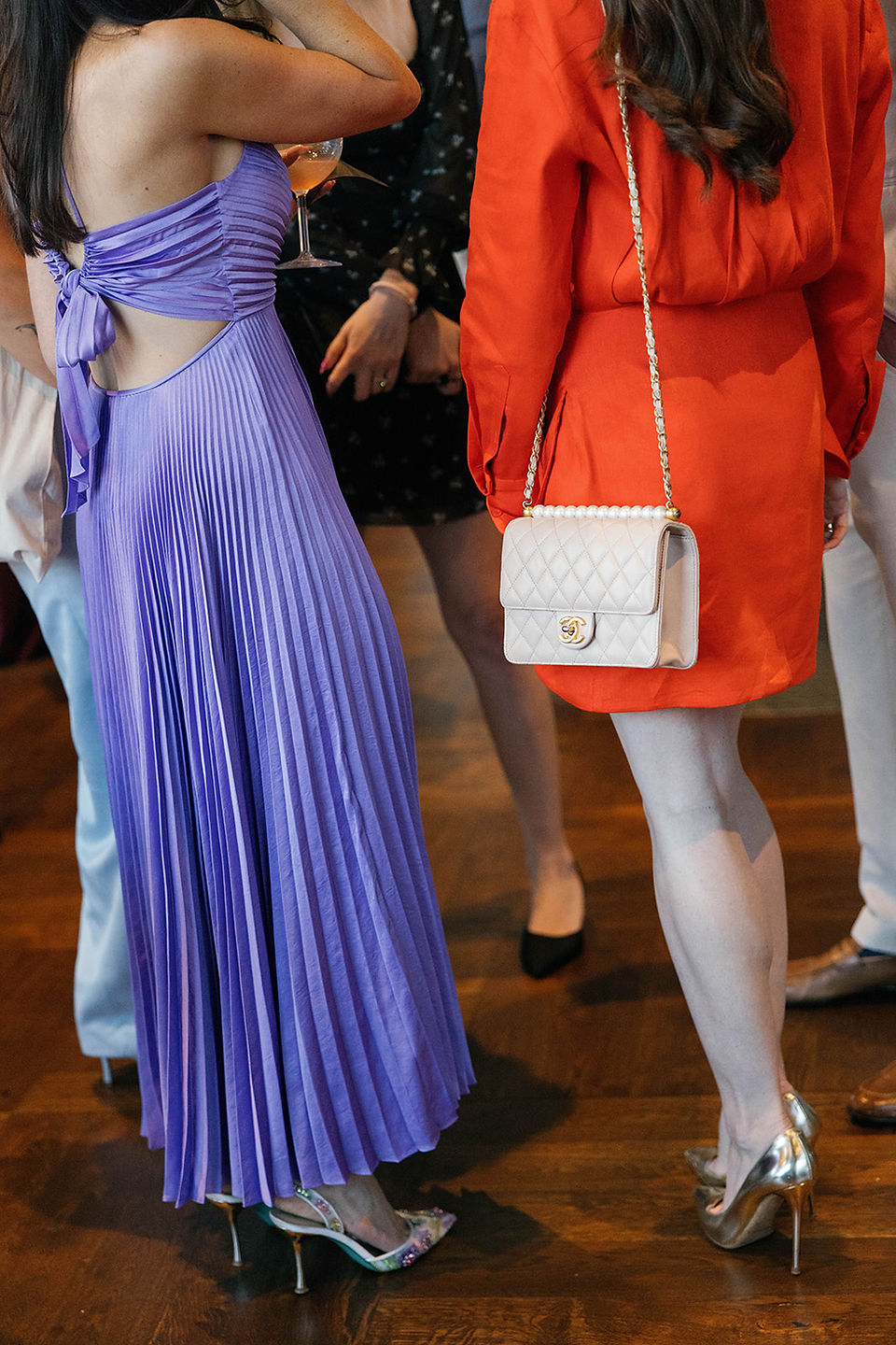 Close-up of bridal shower guest attire featuring a lavender pleated open-back dress and an orange dress with a quilted crossbody bag.