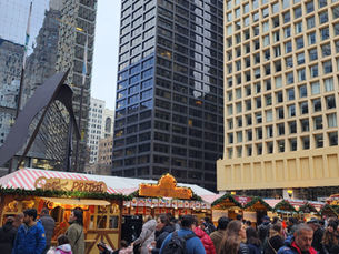 Crowd at an outdoor market with red-and-white tents beneath tall buildings. A man eats food, and signs read Pretzel and Schnitzel.