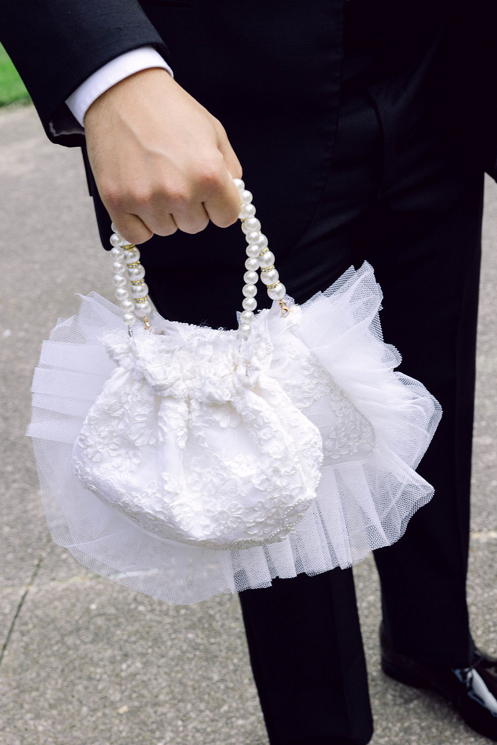 Close-up of a groom holding the bride’s white lace and tulle handbag with pearl handles, highlighting intricate bridal fashion accessories.