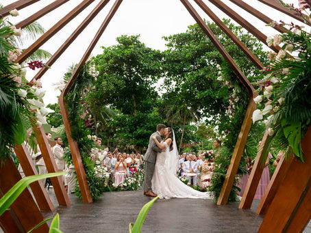 Bride and groom sharing their first kiss at an outdoor destination wedding ceremony in Mexico, framed by a dramatic geometric wooden arch adorned with lush tropical greenery, white roses, and burgundy orchids, as guests celebrate in the background.