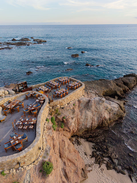 Angled aerial view of the full Cocina del Mar venue at Esperanza Resort, showing the main upper promontory terrace and a curved lower terrace below, both set with teak tables and navy chairs for a wedding dinner, with volcanic rock cliffs, a small sandy cove, and the Sea of Cortez extending to the horizon.