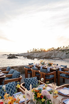 Side-angle view of the Cocina del Mar wedding reception terrace at Esperanza Resort at golden hour, with teak tables and navy cushioned chairs set with colorful summer florals, the volcanic rock cliffs and rocky outcroppings of the Punta Ballena coastline on one side, and the warm glow of a Baja sunset illuminating the palm-lined headland across the water.