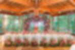Elegant banquet setup with floral arch and vibrant blooms. Neon sign reads "The Robins." Large windows show lush greenery. Cozy ambiance.