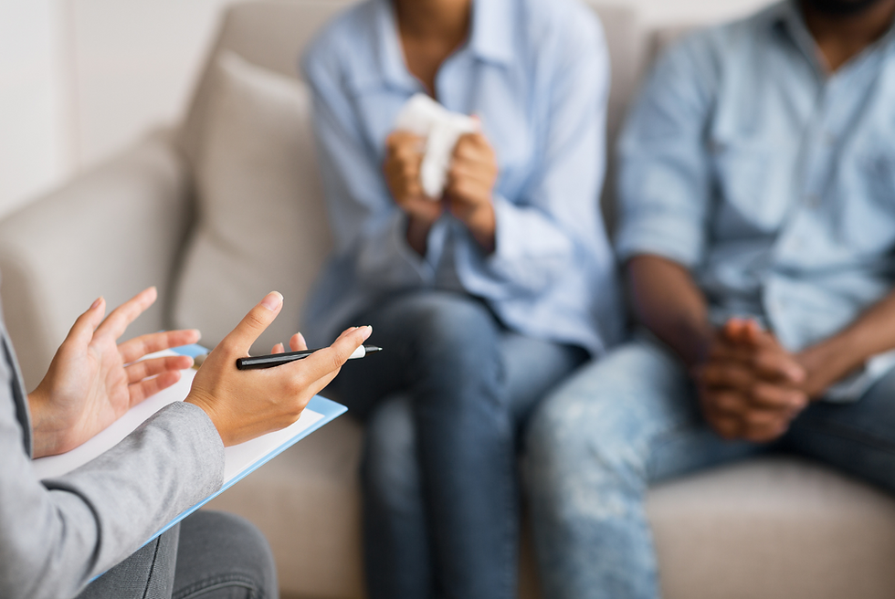 Therapist with a clipboard gestures in discussion. Two people, seated closely, one holding a tissue