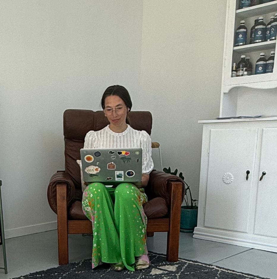 Woman in glasses sits on a brown chair using a sticker-covered laptop in a bright room, wearing a white top and green floral pants.