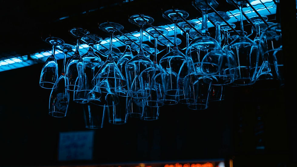 Racks of wine glasses hang in a dimly lit bar, illuminated by neon lights.