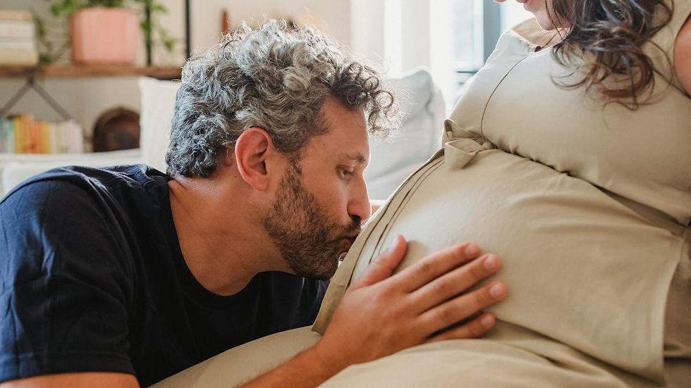 Side view of loving adult husband gently touching and kissing pregnant tummy of crop wife sitting on couch in light apartment