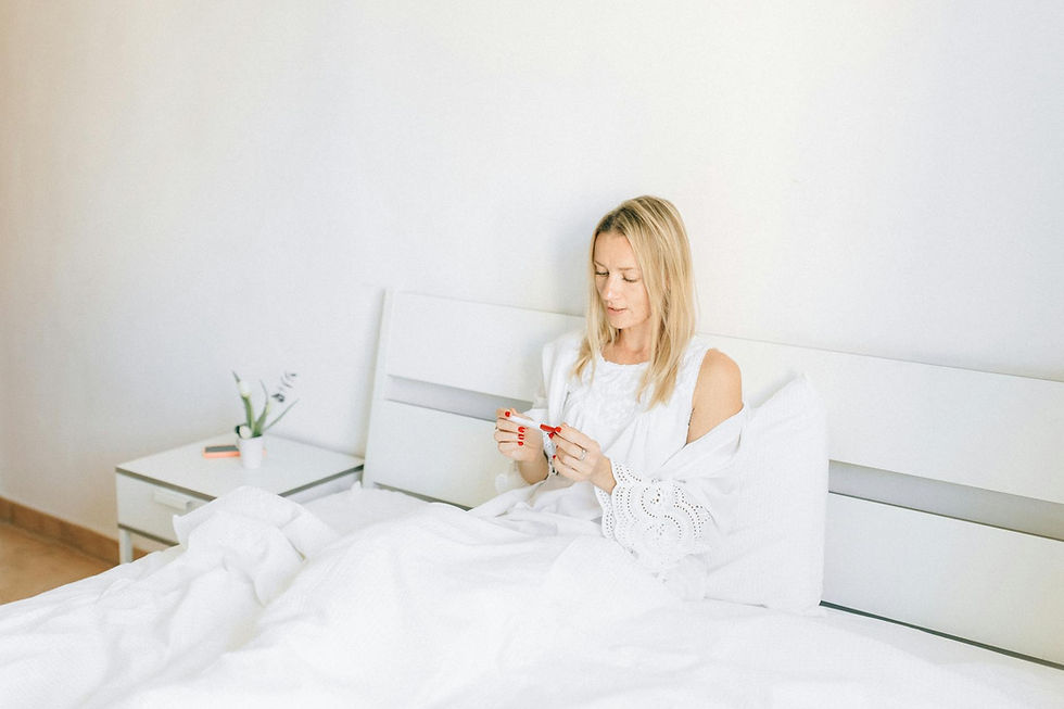 A woman in a white bedroom sits on a bed, holding a thermometer. A potted plant and a notebook are on the bedside table.