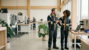 Two manufacturing workers, a white man and a Black woman, converse naturally in a bright, modern production facility. They wear matching dark blue work jackets with high-visibility reflective stripes. The man holds a small metal component while speaking, and the woman listens attentively with one hand resting on a light wood workbench. The airy workspace features large windows, exposed wood beams, lush potted plants, and modern machinery with other employees working in the softly blurred background.