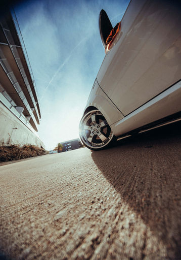 Low-angle view of a white car with silver alloys on textured concrete, featuring modern flats and a blue sky.