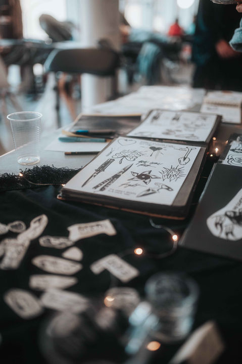 Detailed tattoo flash sketches on a sketchbook at an art event exhibition, featuring black ink and warm bokeh lighting.