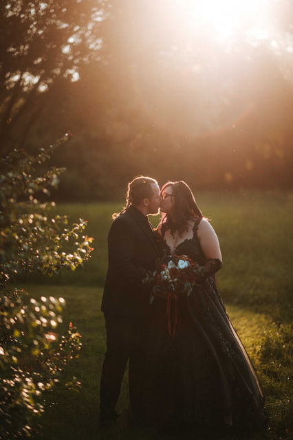 Couple kissing in golden sunset light for svatby; bride in a black lace dress holds a red and white floral bouquet.