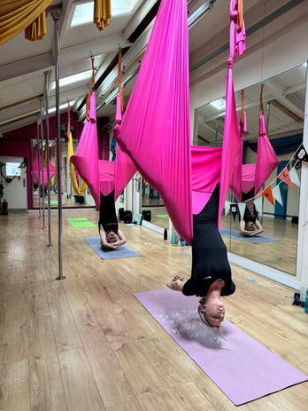 Women perform aerial yoga inversion in pink hammock within bright studio.