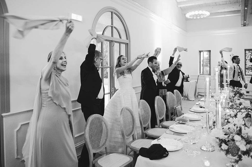 Black-and-white wedding reception with guests and bride waving napkins beside a long floral banquet table, cheering joyfully
