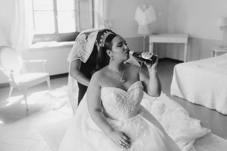 Funny black and white portrait of bride drinking beer while make up artist set veil