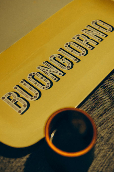 cup of coffee beside a yellow placemat written good morning in Italian