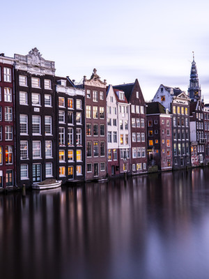 A serene evening settles over Amsterdam’s iconic canal houses, their warm lights dancing on the calm water below.