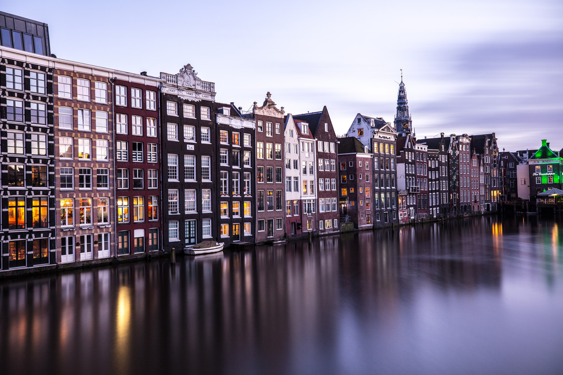 A serene evening settles over Amsterdam’s iconic canal houses, their warm lights dancing on the calm water below.