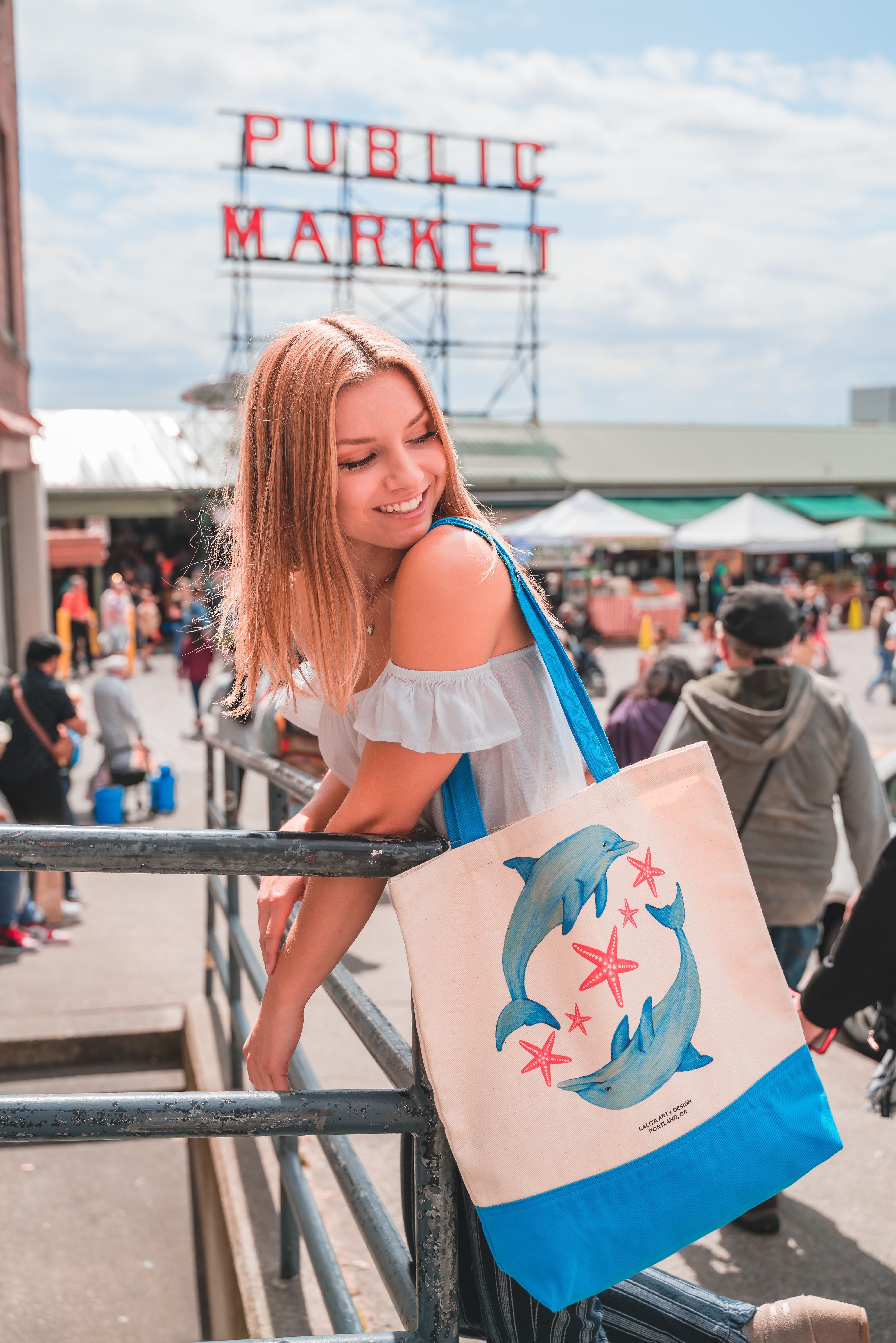 Young woman smiling, holding tote bag at Pike Place Market with MARKET sign.