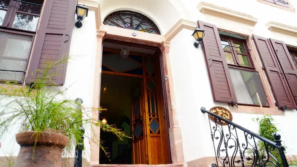 Beyaz dış cephe ve ahşap panjurlu Zehra Teyze'nin Evi'nin açık kapısı. Yanında saksı bitkileri var. Ferforje korkuluğun yanında duvarın üzerinde otelin tabelası asılı.