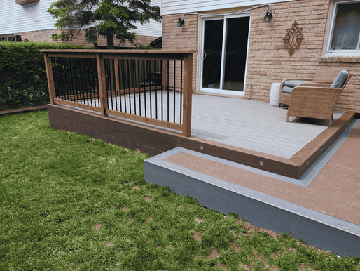 Wooden deck with a wicker chair, bordered by a black-railed fence. Green lawn and brick wall in the background, creating a serene setting.