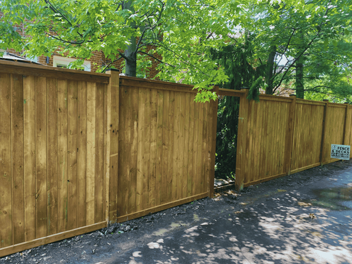 Wooden fence under green trees with sunlight filtering through. A sign reads "Fence & Decks" with a phone number. Peaceful setting.