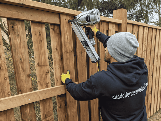 Man in hoodie and gloves uses a nail gun on a wooden fence outdoors. Text on hoodie: citadelfenceanddeck.ca. Overcast day.