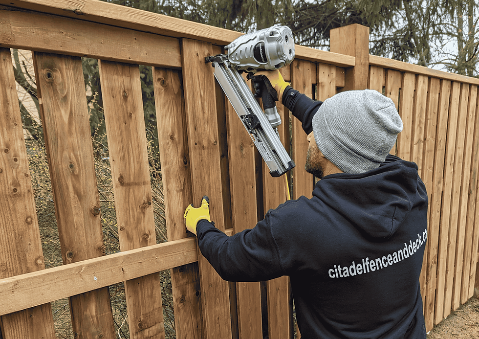 Man in hoodie and gloves uses a nail gun on a wooden fence outdoors. Text on hoodie: citadelfenceanddeck.ca. Overcast day.