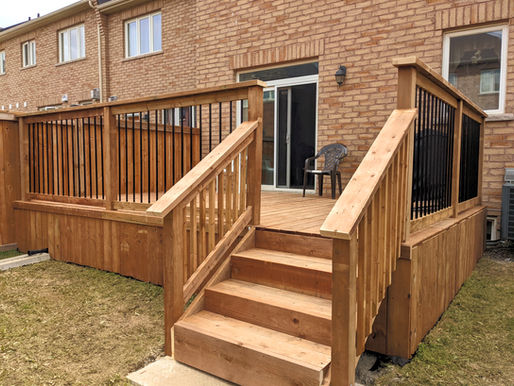 Wooden deck with black railings on a brick house. Steps lead up. A plastic chair is visible. Overcast sky gives a calm mood.