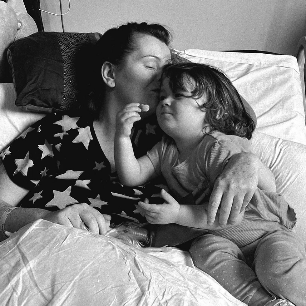 A black and white image of Leanne holding her young child in bed, she's kissing the child on the head. 