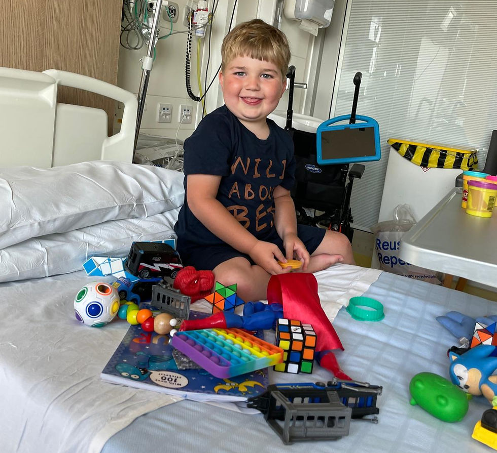 Rowan in hospital, surrounded by toys. He's smiling on the bed.