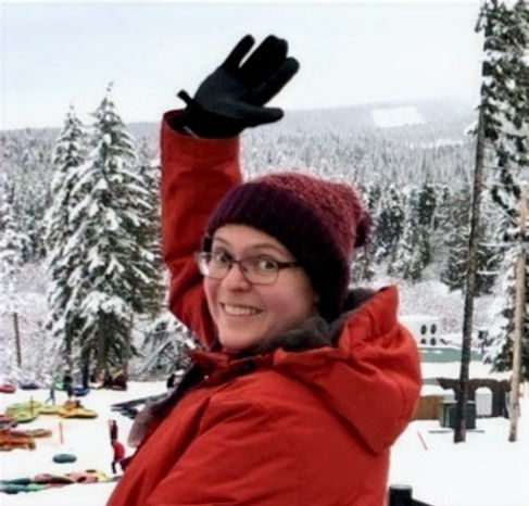 A picture of Dragonfly Consultant's Principal, Shelley Beaulieu, in an orange heavy coat looking over her shoulder, waving at the camera. She is on a snowy, tree-covered hill.