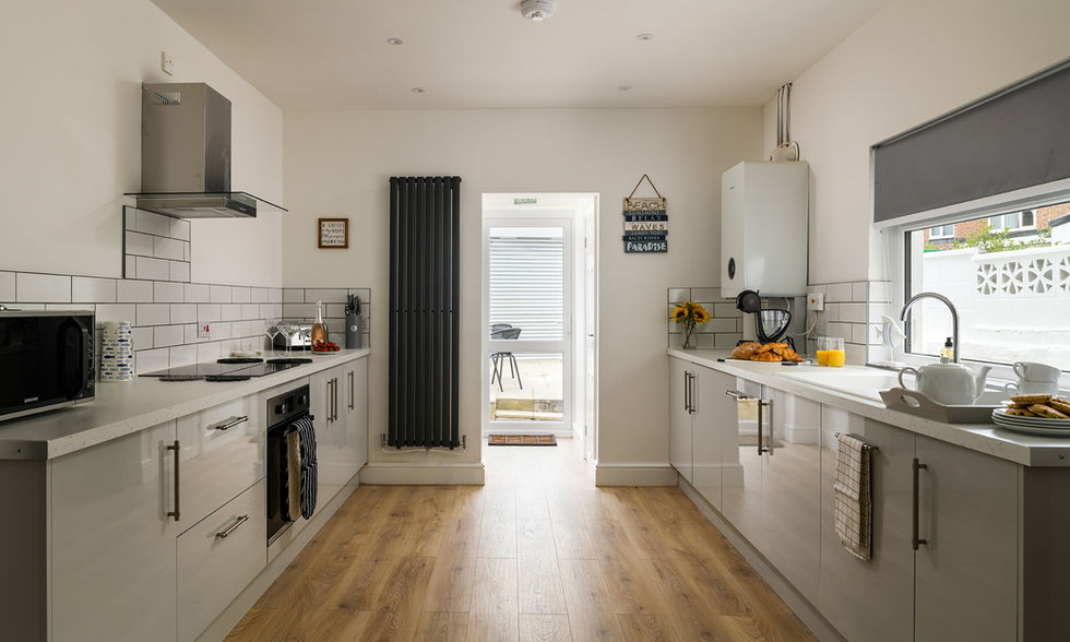 A long kitchen with brown wooden flooring and white kitchen cupboards and a back door and a long grey radiator on the wall.