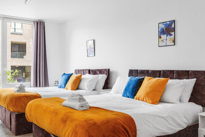 Two beds in a hotel room with orange and blue pillows.