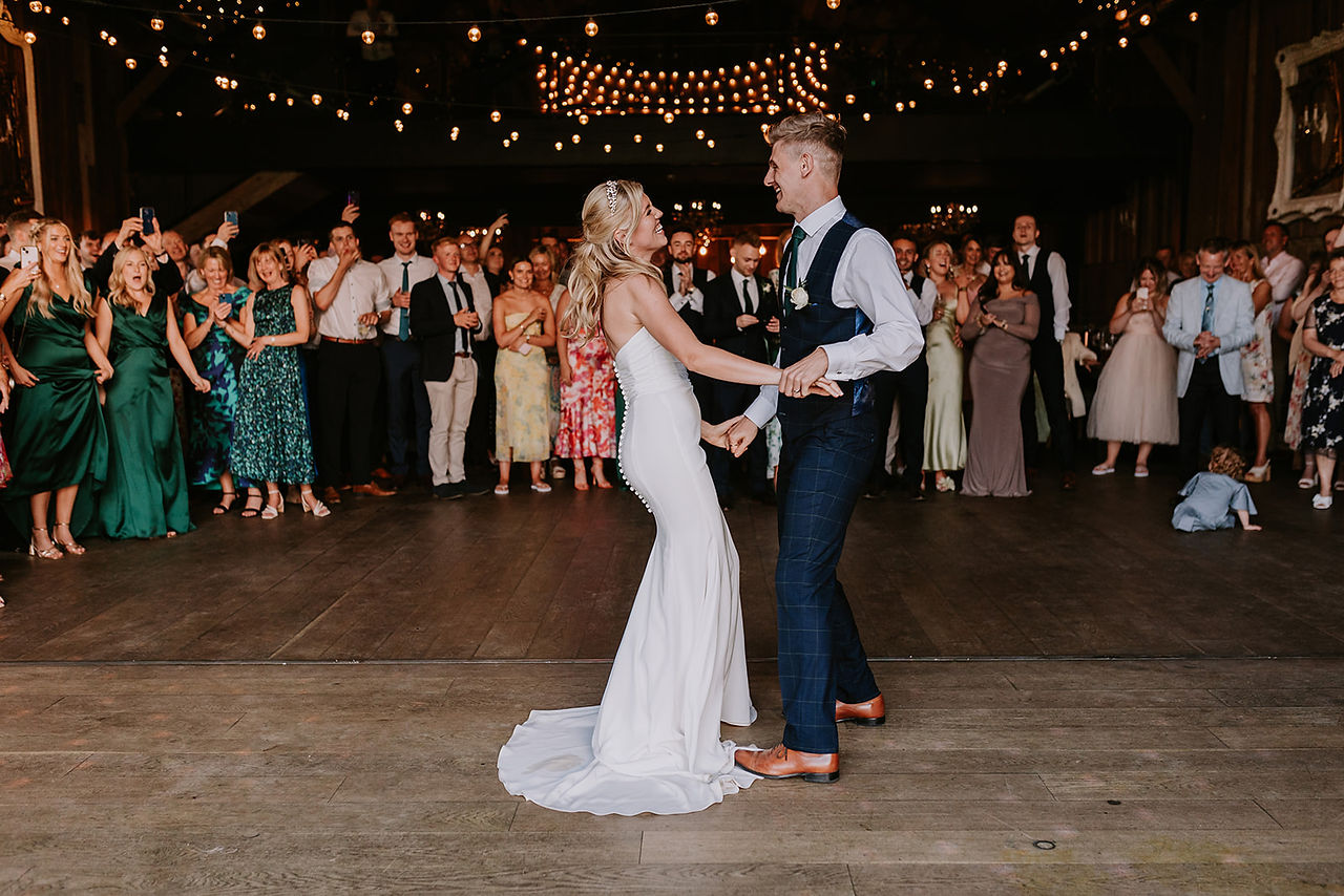 The bride and groom are dancing and smiling at each other during their first dance. The guests are all watching and clapping and smiling.