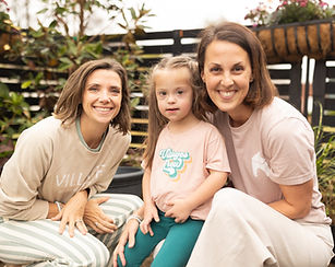 a photo of two adults sitting with a young girl with Down syndrome, leaning in closely and smiling together at the camera