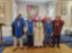 Brothers of the Order in blue and red regalia standing with a priest in a room at Ely House, beneath a painting and the Order's banner.