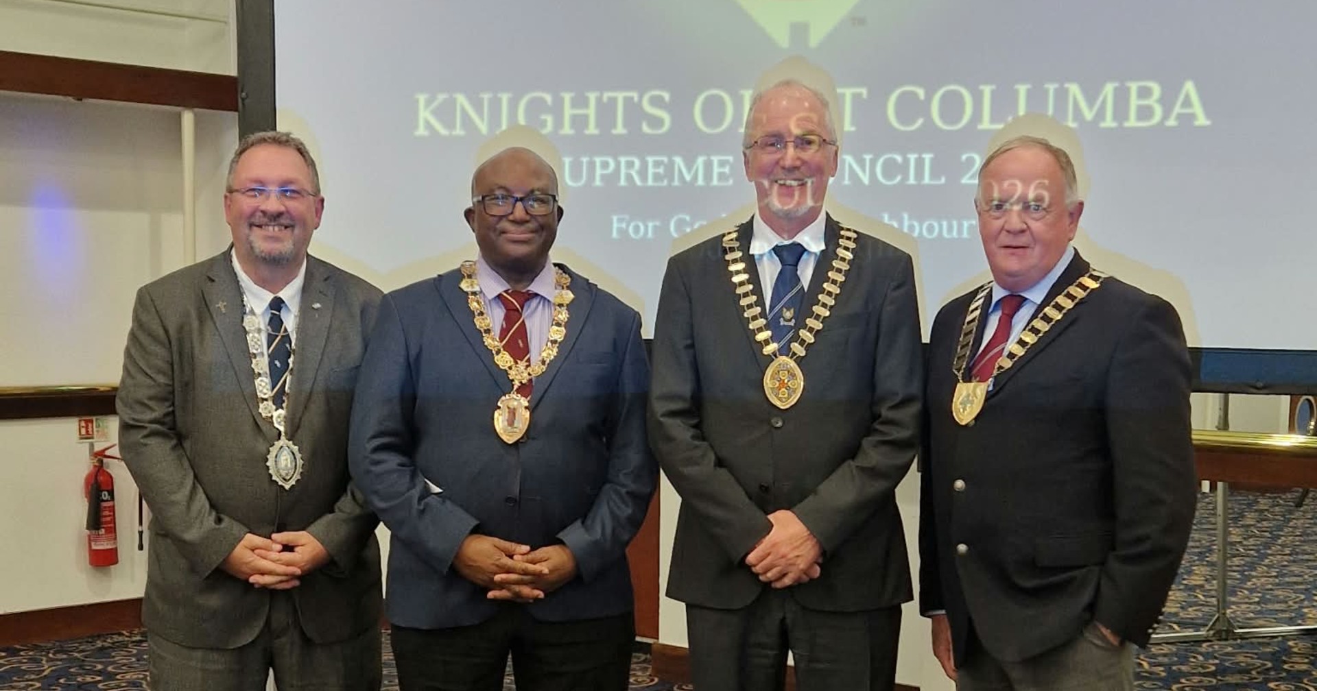 Four men in suits stand side by side on a patterned blue and gold carpet, each wearing a chain of office with a large medallion, their hands clasped in front of them. Behind them a screen shows the words Knights of St Columba, Supreme Council 2026.