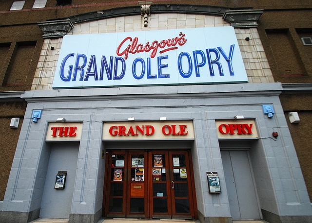 Glasgow's Grand Ole Opry entrance with red doors, posters displayed. Blue and red signage contrasts with the brown brick building facade.