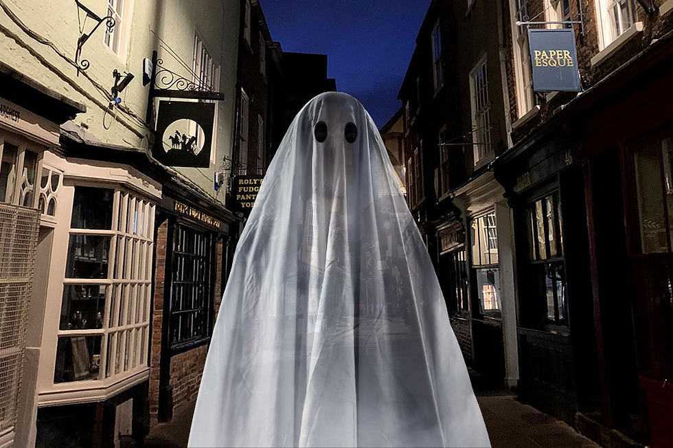 A ghostly figure in a white sheet with cut-out eyes stands in a dimly lit alley with historic buildings and shop signs at night.