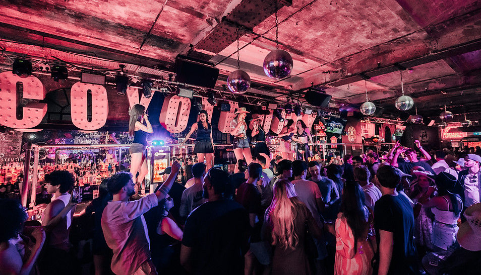 Crowded bar scene with people dancing. Women perform on a bar top under pink lighting. Disco balls and large "COYOTE" sign hang above. Mood lively.
