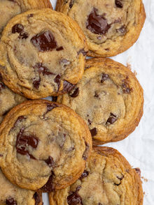 Chocolate chunk cookies are layers on top of crinkled white parchment paper with light brown centers and golden edges and melty chocolate chunks and chips throughout the cookies