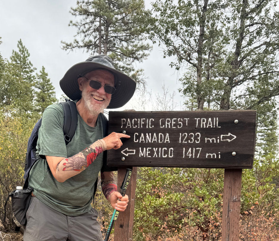 a male hiker smiles, and while standing at a wooden PCT sign, points to the distance noted regarding Canada..