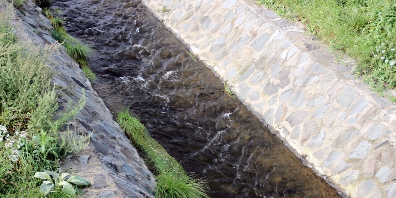 Narrow stone-lined drainage with flowing dark water, bordered by green grass and foliage, showcasing professional drainage services in Kingswell.